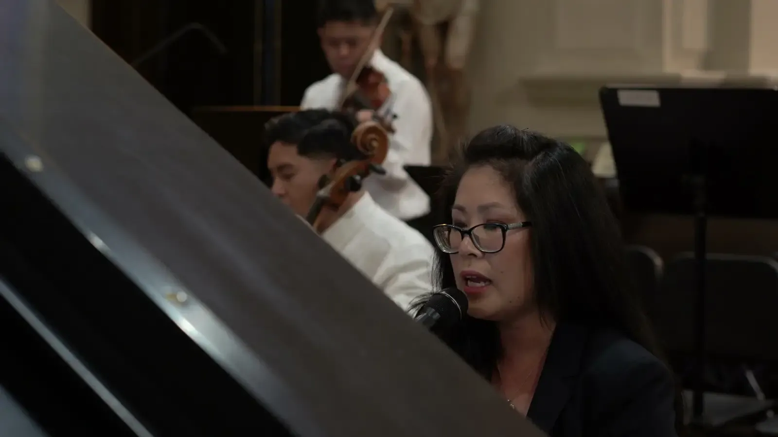 Anhthu singing at the piano during a Mass, a cello and violin behind her in a warm-lit church